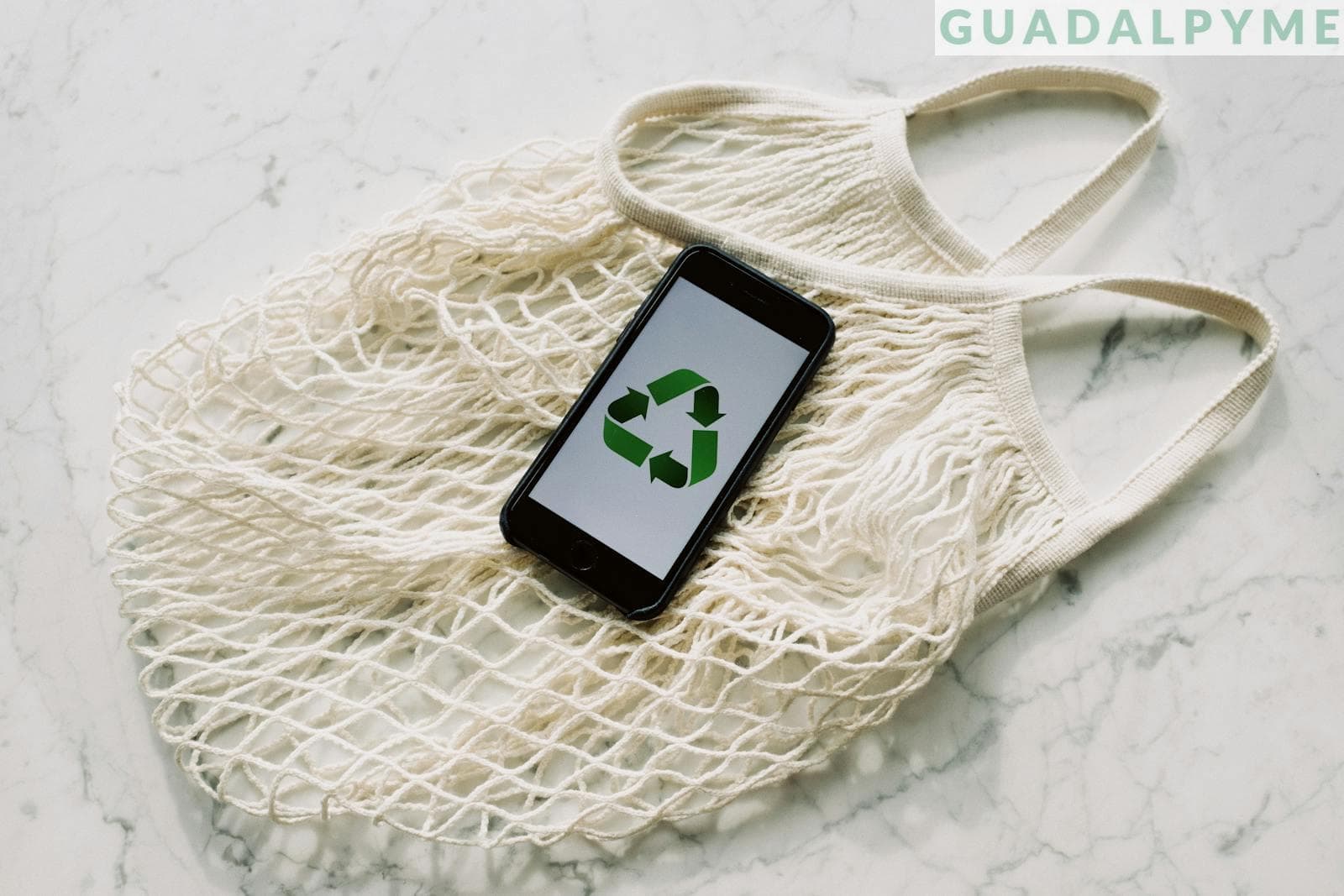 Overhead of smartphone with simple recycling sign on screen placed on white eco friendly mesh bag on marble table in room